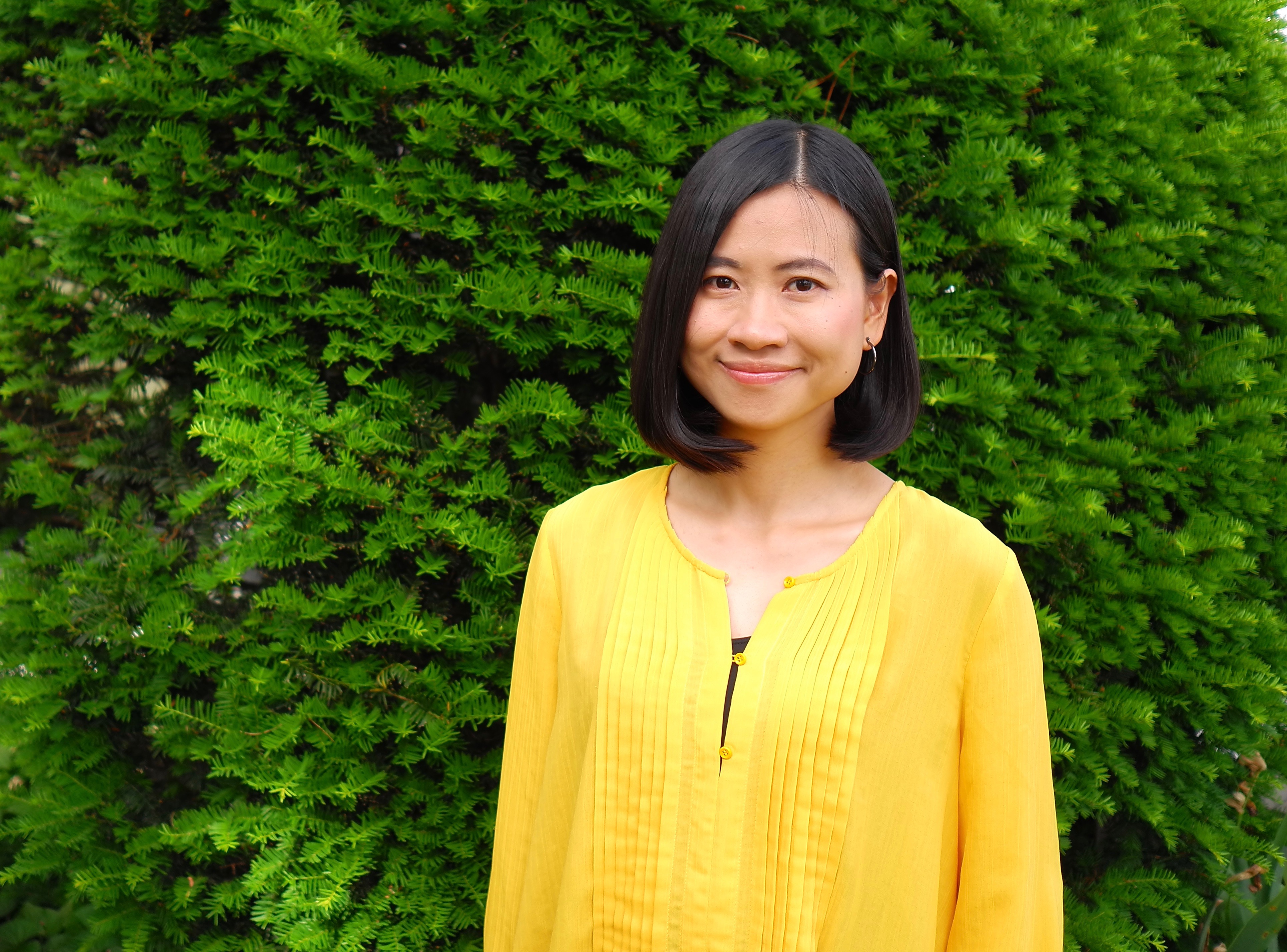 Graduate student Kongjing Xing wears a bright yellow shirt standing in front of a leafy, green tree