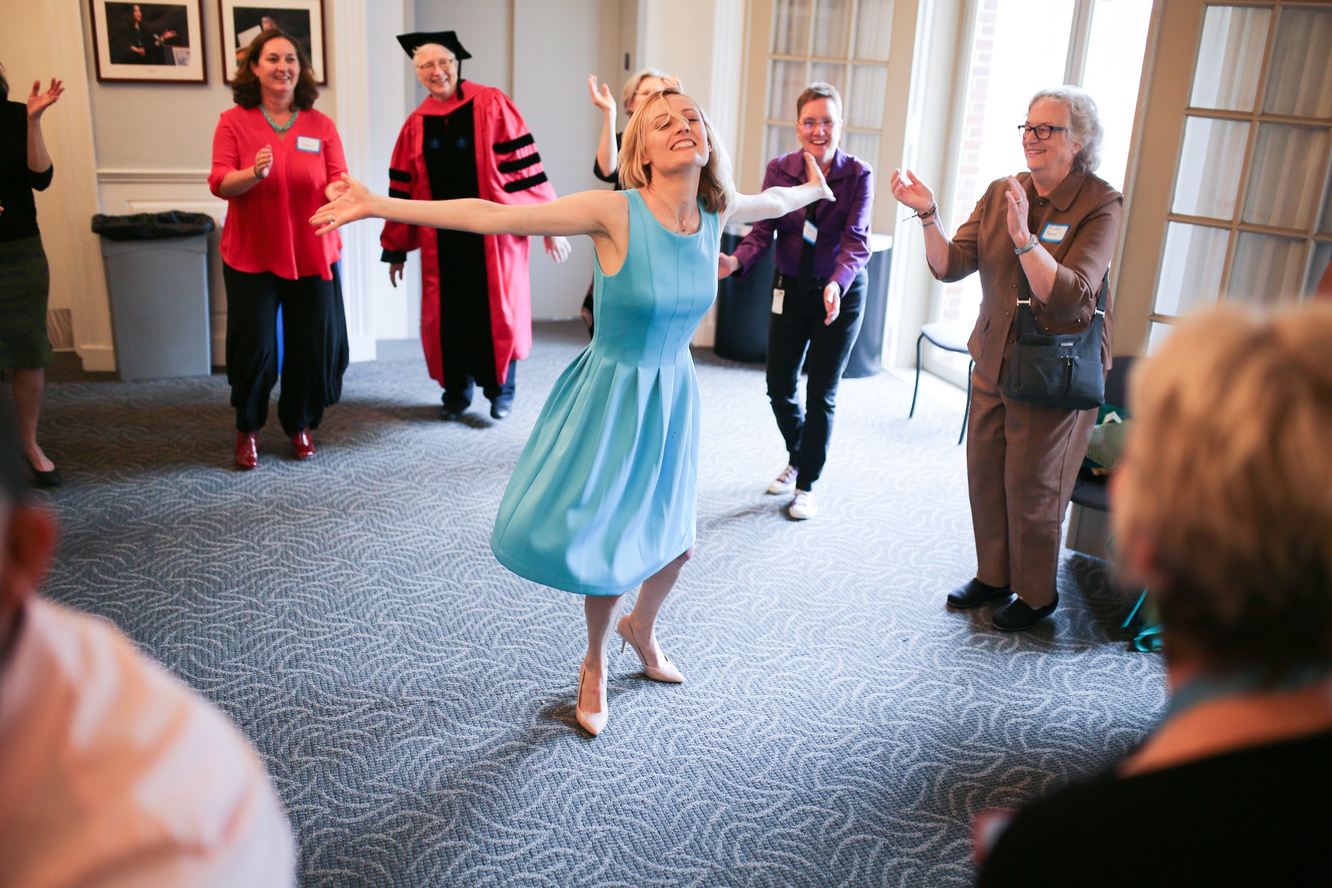 Photograph of a young woman in a blue dress dancing in a circle of people inside in a room with a carpet.