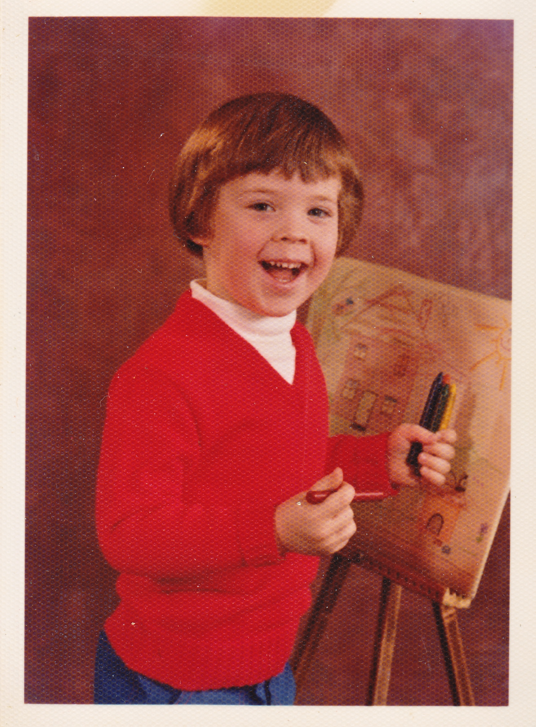 Keith MacLelland as a kid smiling at the camera with an easel and artwork behind him and crayons in his hand.