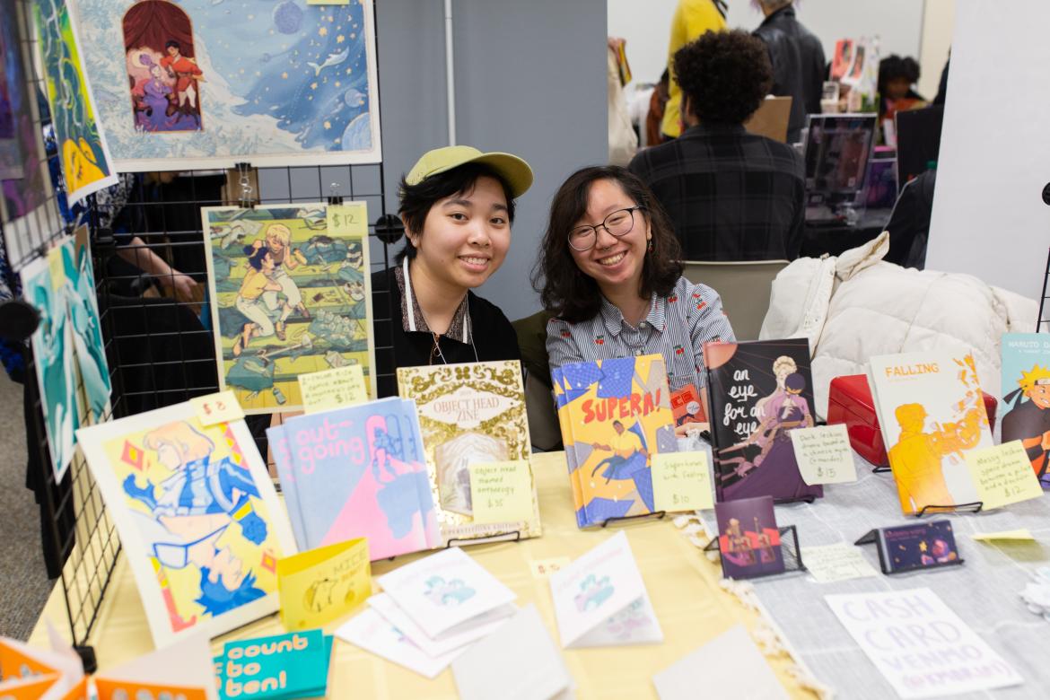 2 artists behind a table at a comic book convention with books and papers in front of them