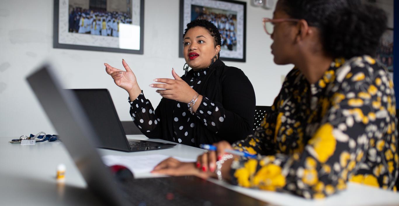  Two women are engaged in a professional discussion at a conference table with open laptops and documents. The woman in the center, wearing a black polka-dotted blouse and gesturing with her hands, is speaking confidently. The woman on the right, wearing a floral-patterned outfit, listens attentively. The setting appears to be an office or academic environment, with framed photos on the wall in the background.