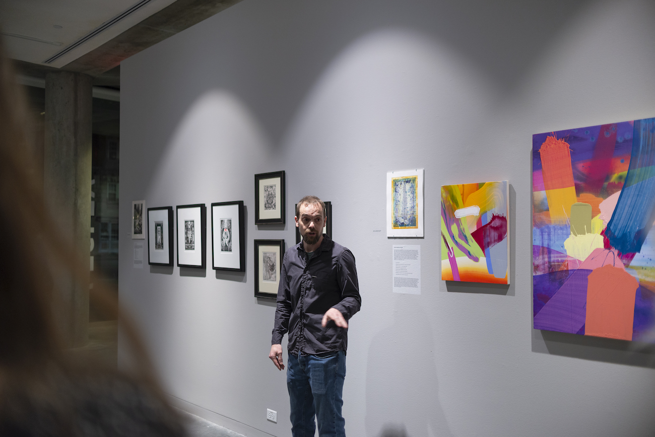 Printmaker Adam Amara discussing his art pieces against a grey wall and a row of picture frames
