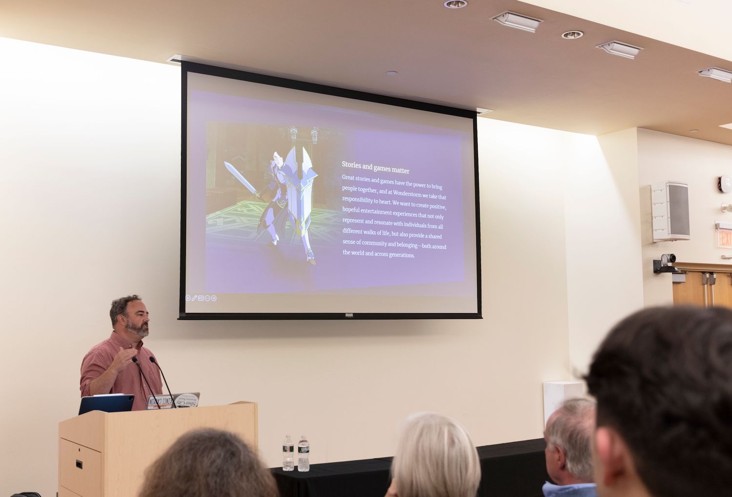 Justin Richmond stands behind a podium next to a screen projector giving a presentation to a lecture hall of people