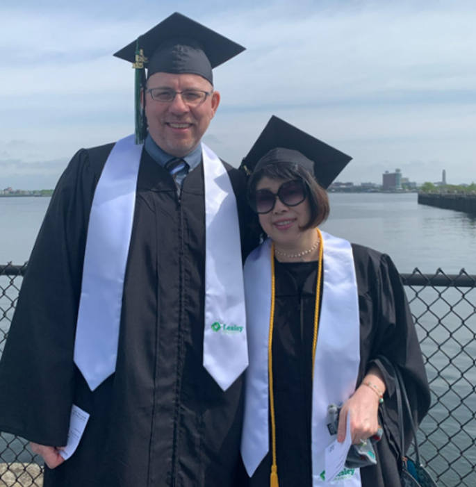 Lesley graduates Hiroko and John Warford stand in their regalia in front of an ocean view