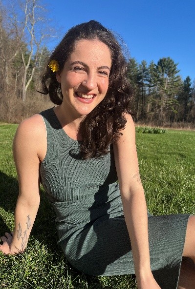 Individual sits in a field in a green dress, facing the camera and smiling.