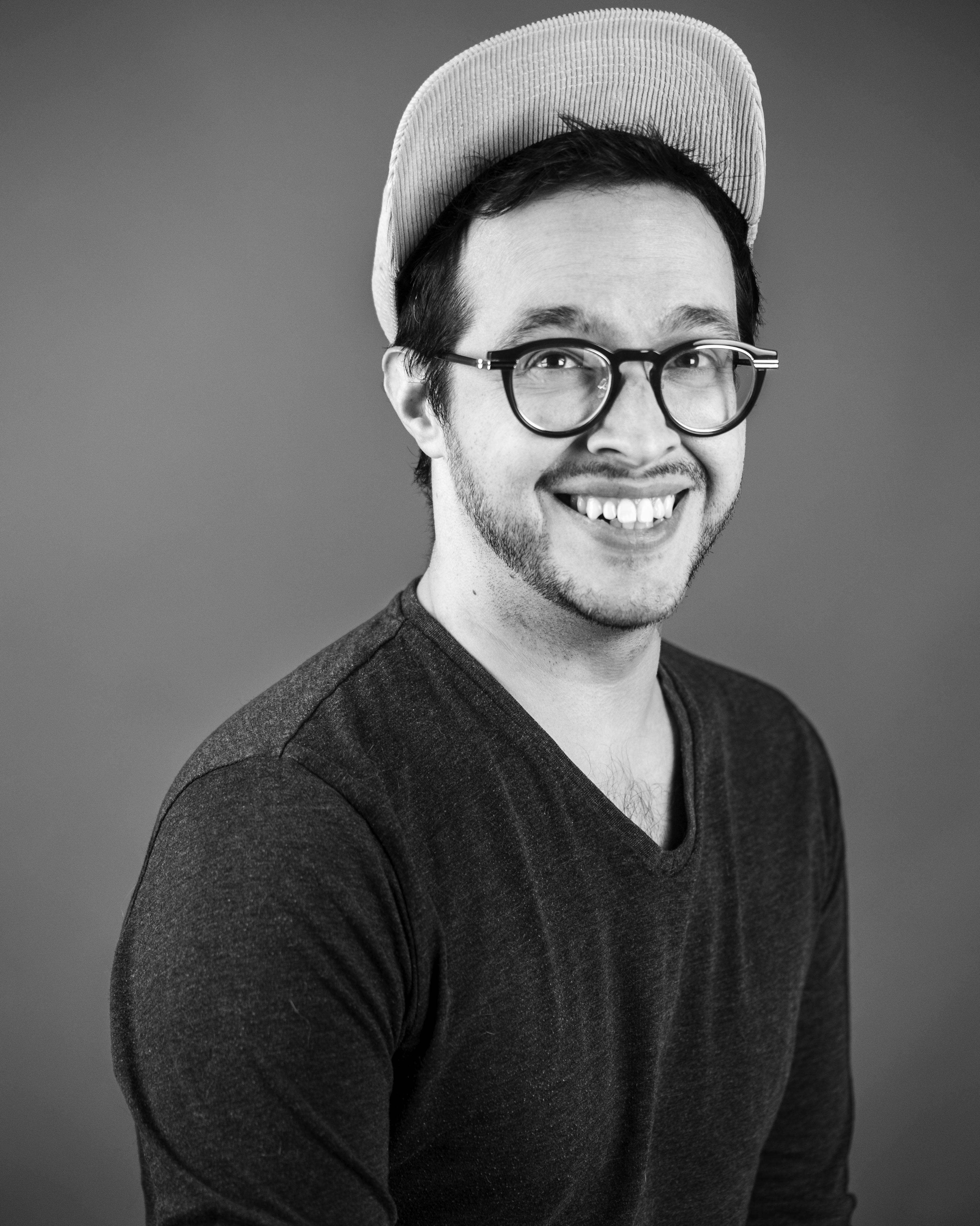 Tynan smiling wearing glasses and a hat in black and white