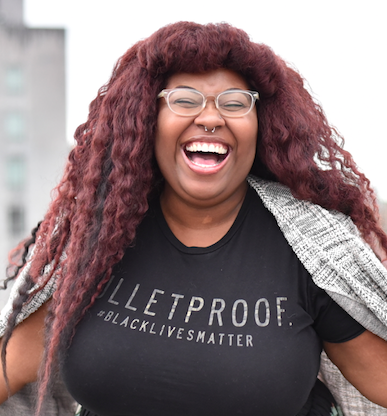 A photo of Daunasia Yancey. She is a Black women with curly red hair and glasses. She is wearing a black t shirt that says "BULLETPROOF #BLACKLIVESMATTER" with a grey cardigan. 