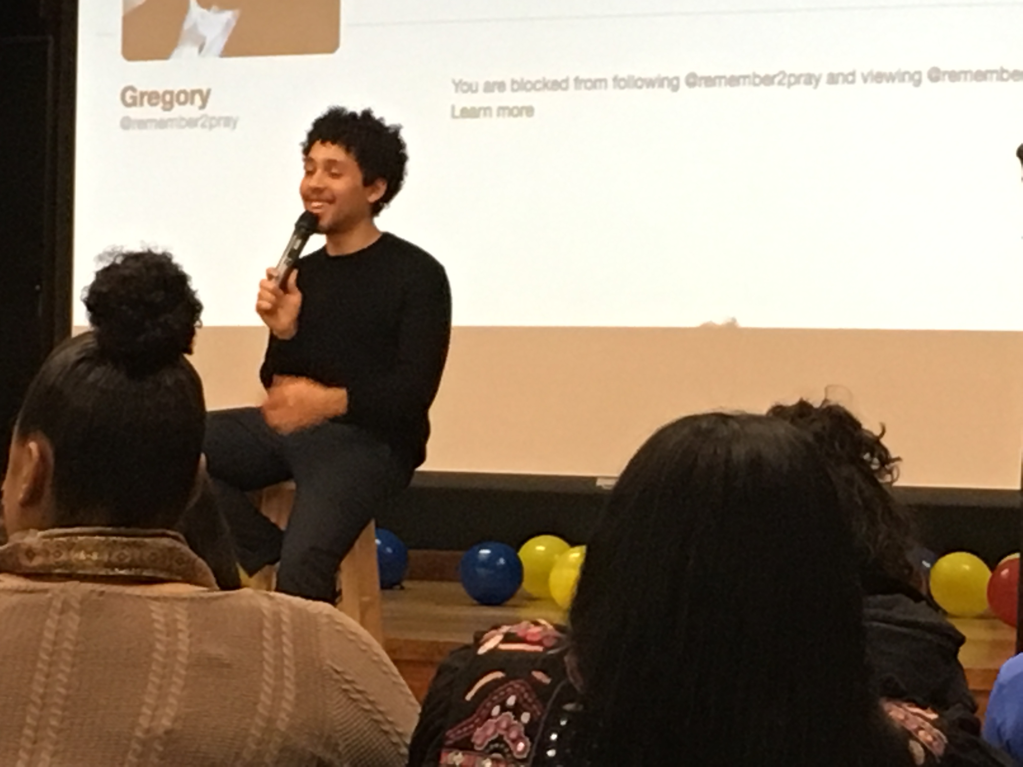 Jaboukie Young-White is perched on a stool in front of a screen, holding a microphone