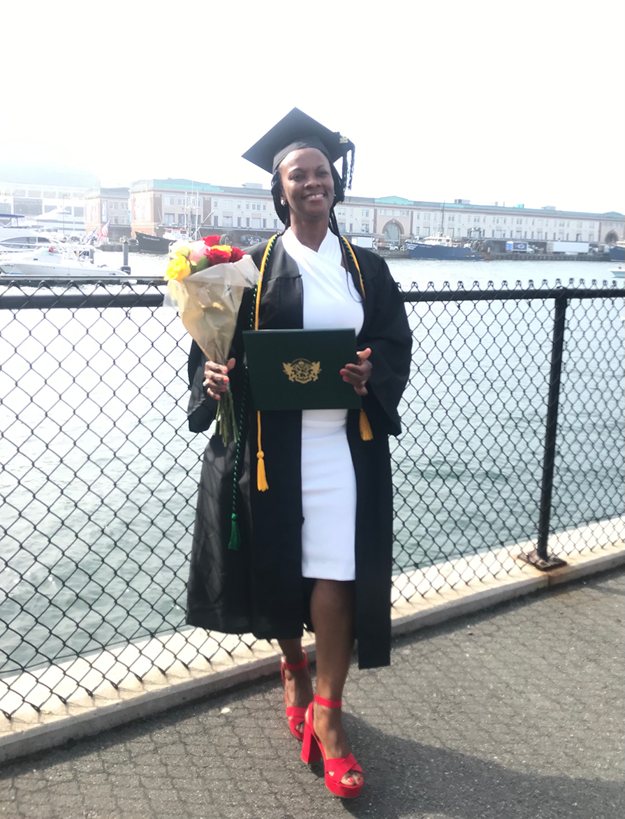 Julette Moore posing for a picture on the waterfront of Leader Bank Pavillion at the 2022 Commencement Afternoon Ceremony. She is wearing a cap and gown and is holding her degree in her left hand and flowers in her right hand.