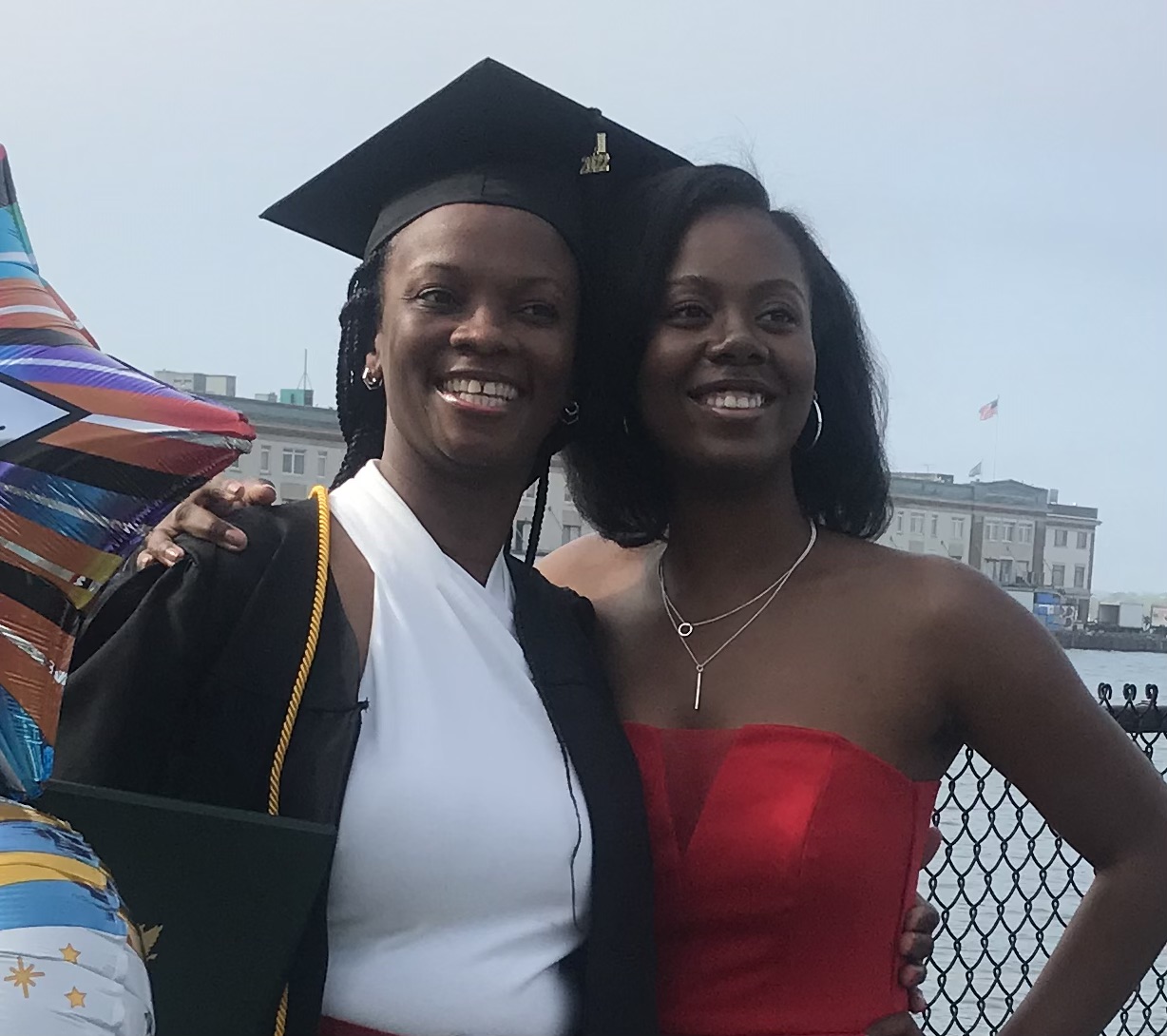 Julette Moore (left) and her daughter Rheanna (right) posing for a picture by the waterfront at the Leader Bank Pavillion at the 2022 Commencement Afternoon Ceremony. Julette is wearing a cap and gown, a white dress, and a yellow cord around her neck. Rheanna is wearing a red top. There are balloons in the background to the left of Julette.