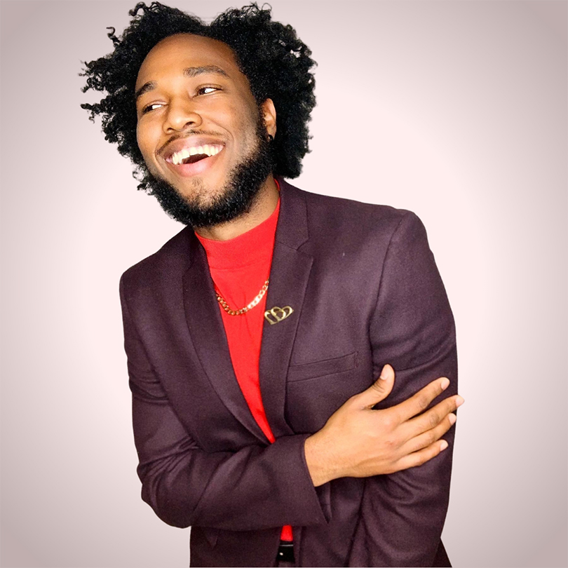 A photo of Lamb Rahming. He is a Black man with curly hair. He is wearing a purple blazer over a red sweater.