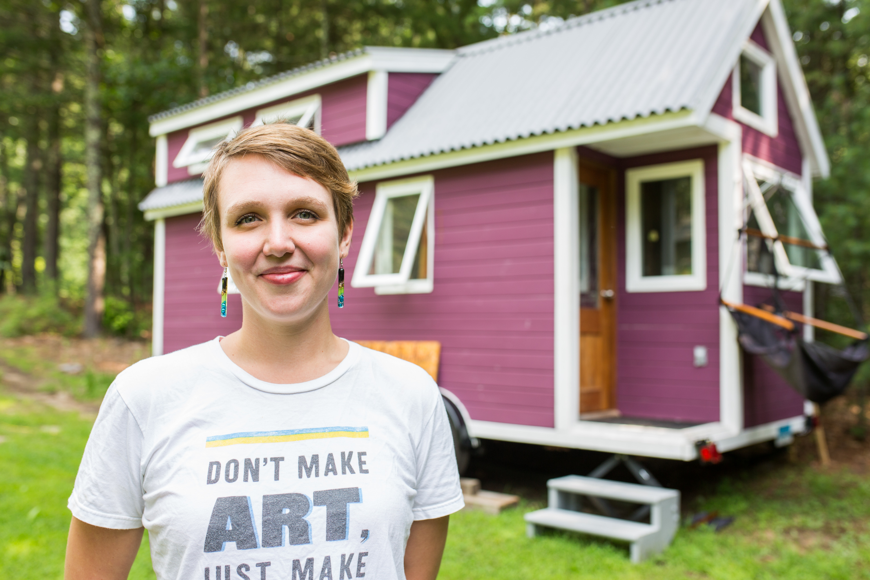 Miranda Aisling stands outside of her purple tiny house nestled against a backdrop of trees.