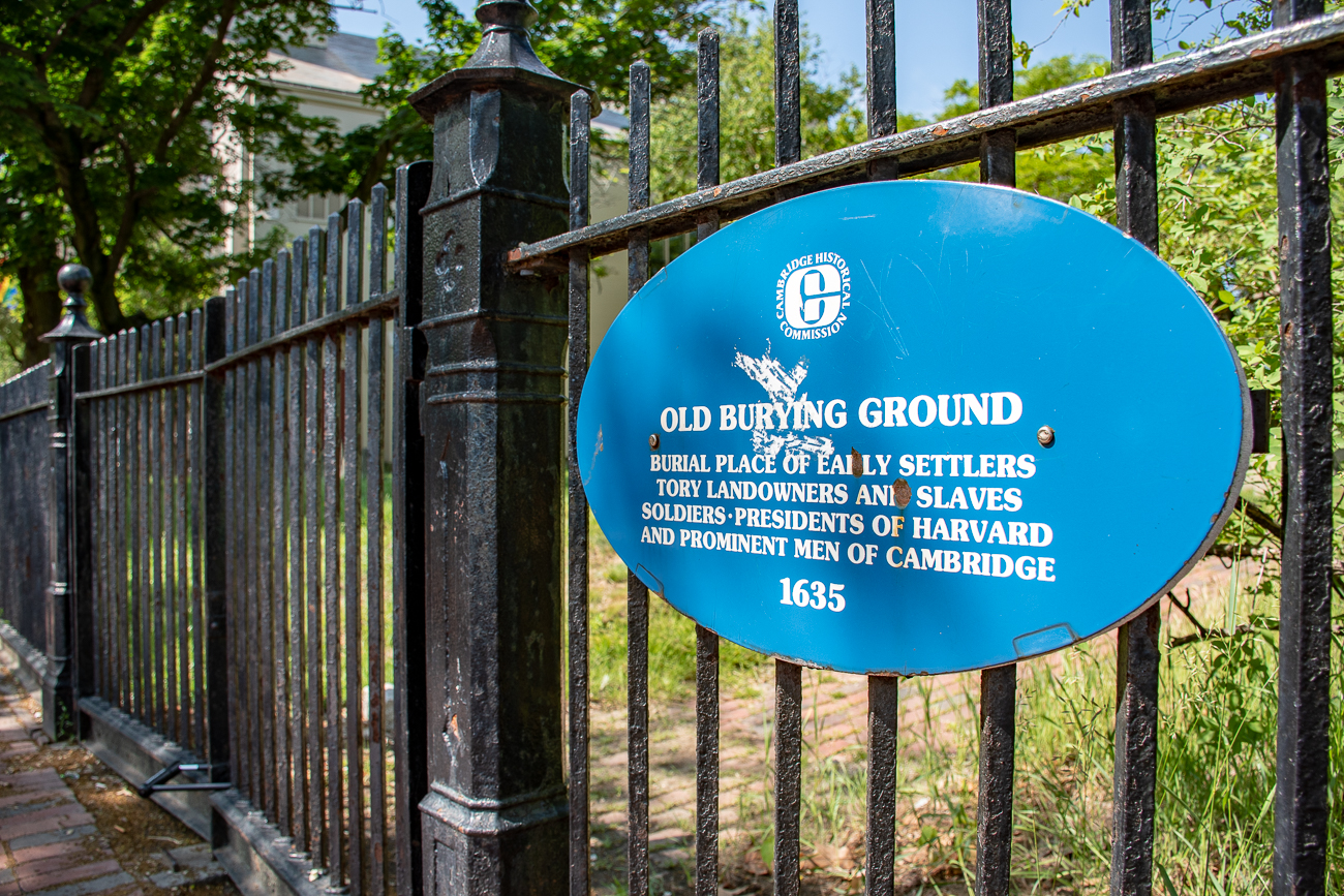 Blue plaque on the wrought iron cemetery fence reads: Old Burying Ground. Burial place of early settlers, tory landowners and slaves, soldiers, presidents of Harvard and prominent men of Cambridge.