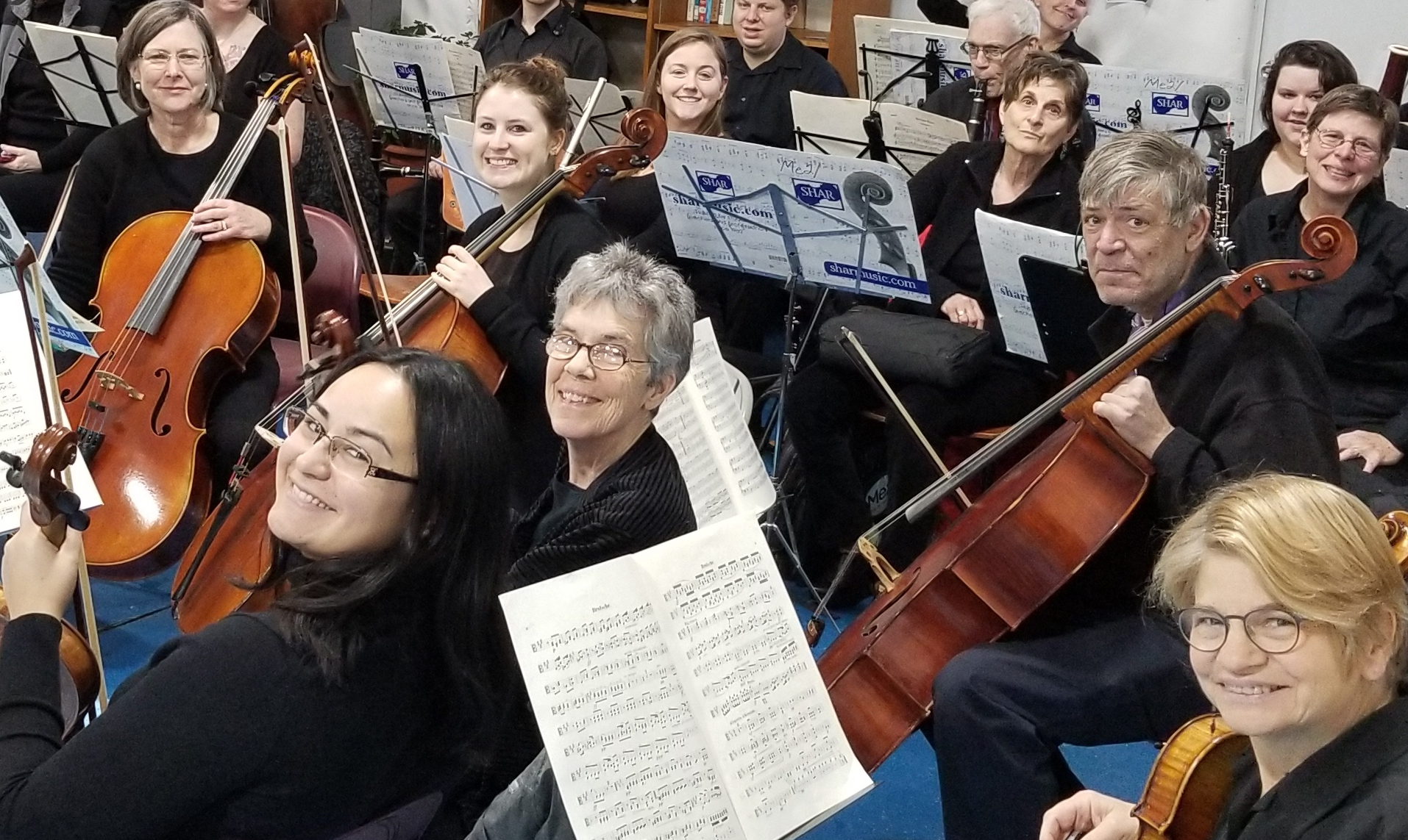 An orchestra seated in with men and women holding instruments including violins, cellos and clarinets.