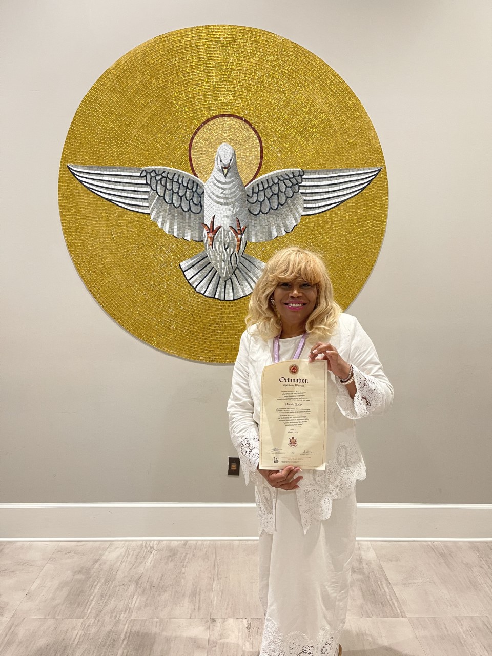 Pamela Kelly in front of a mural of a dove surrounded by gold. Holding a scroll.