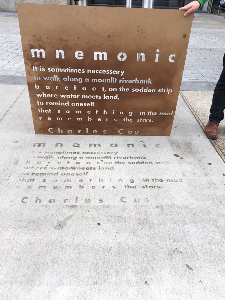 "Mnemonic" by Charles Coe is painted on a sidewalk in Boston. Someone holds the stencil.