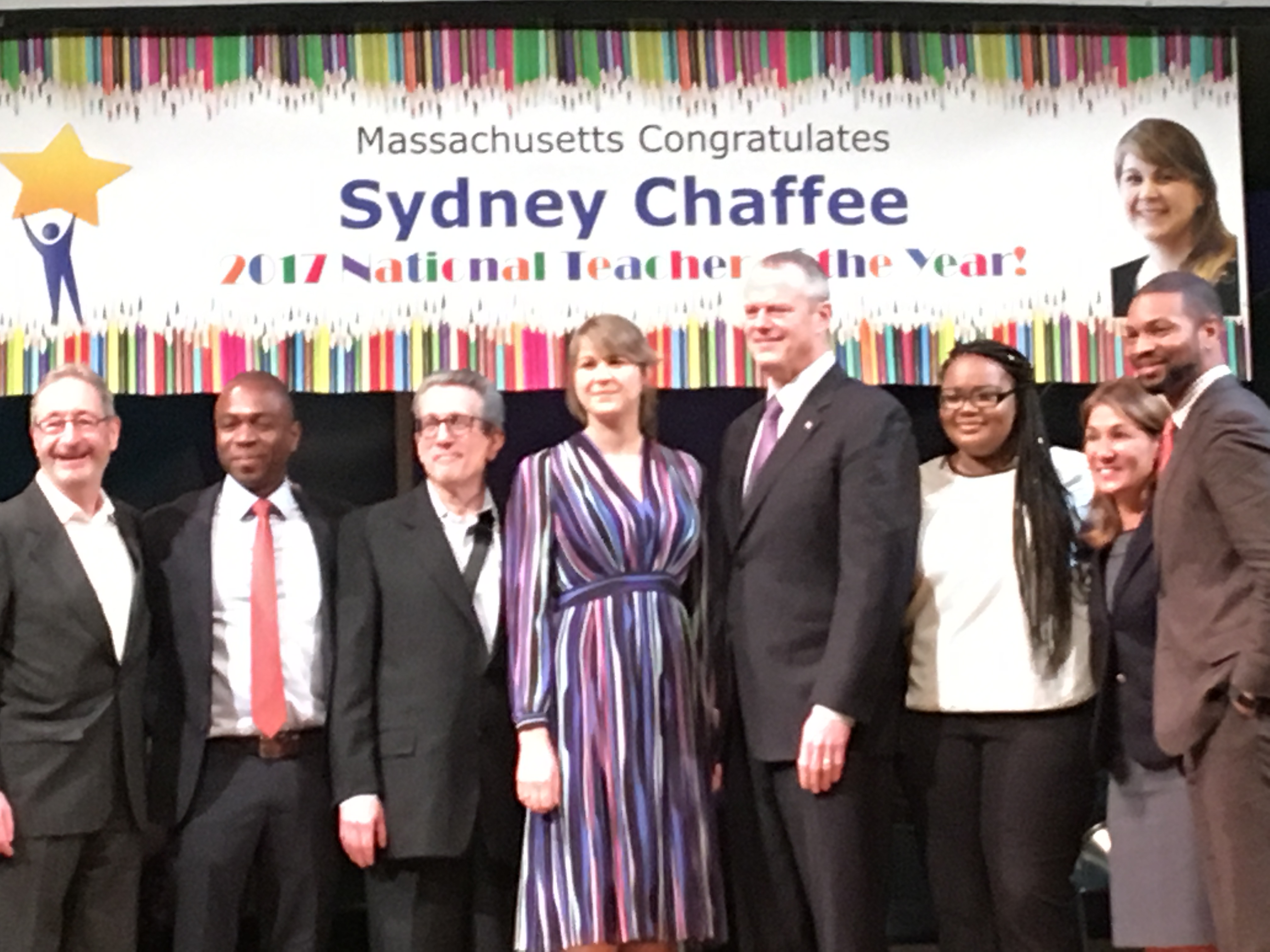 Sydney Chaffee stands on stage with Gov. Charlie Baker, colleagues and students at her ceremony. Behind her is a sign that reads Massachusetts Congratulates Sydney Chaffee.