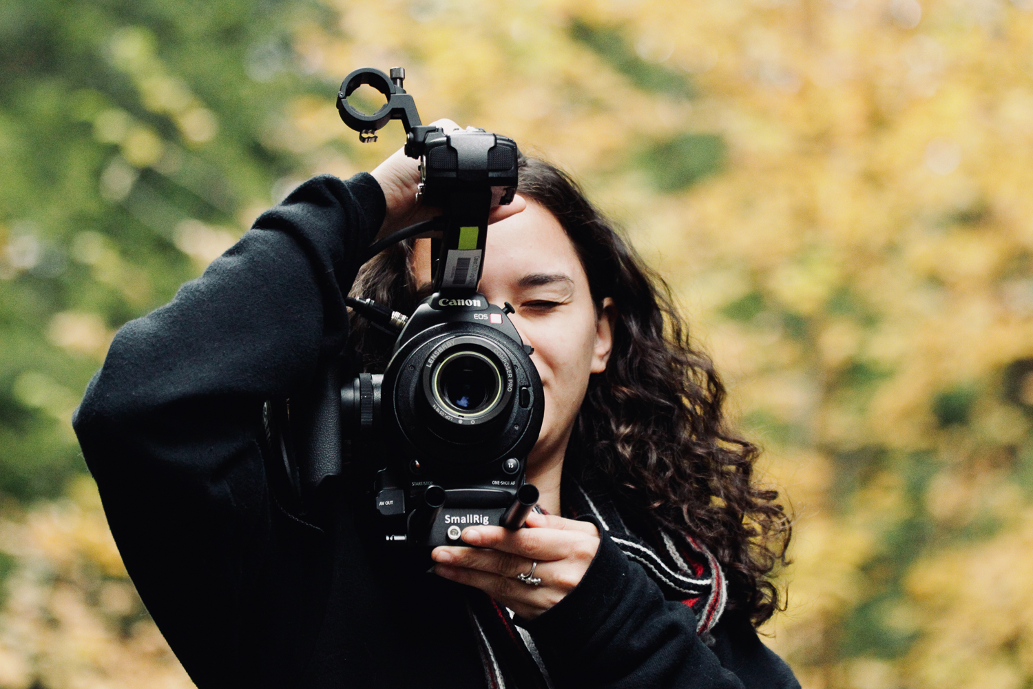Emily Barone holding a large camera in the woods with yellow autumn leaves in background