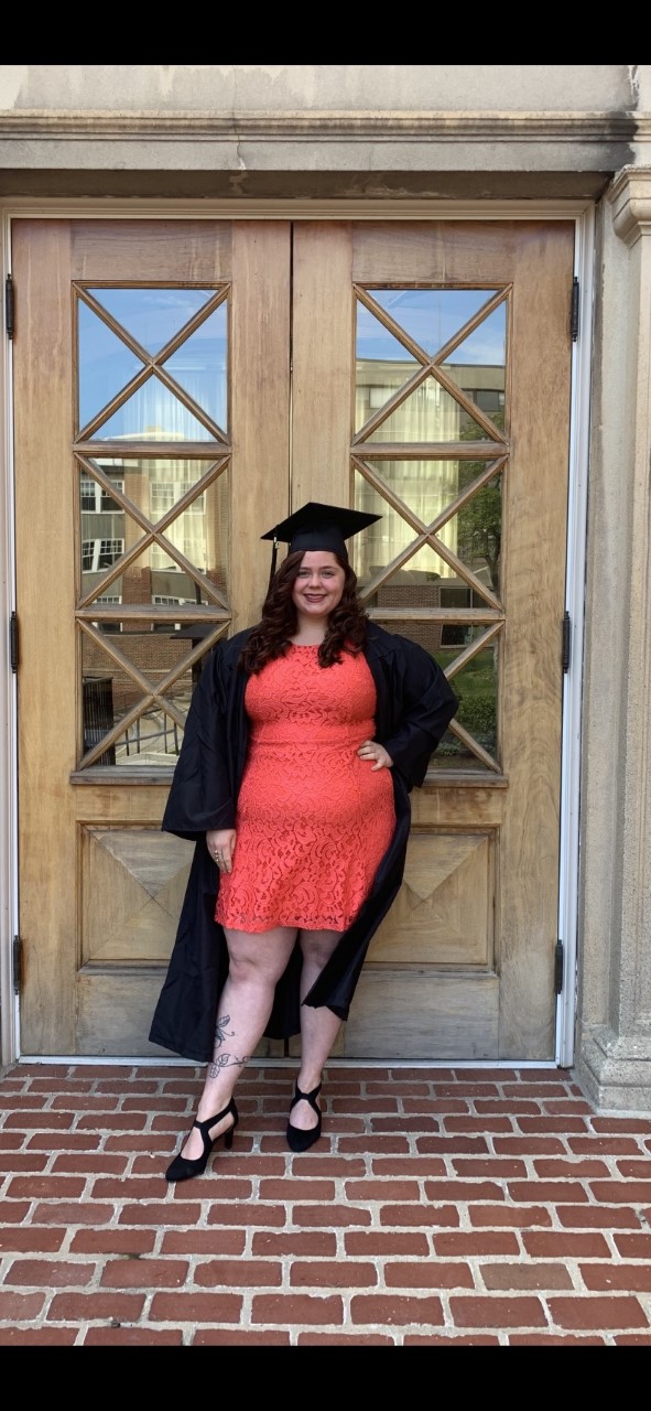 student zandra matthews standing in front of a door wearing a graduation cap and gown