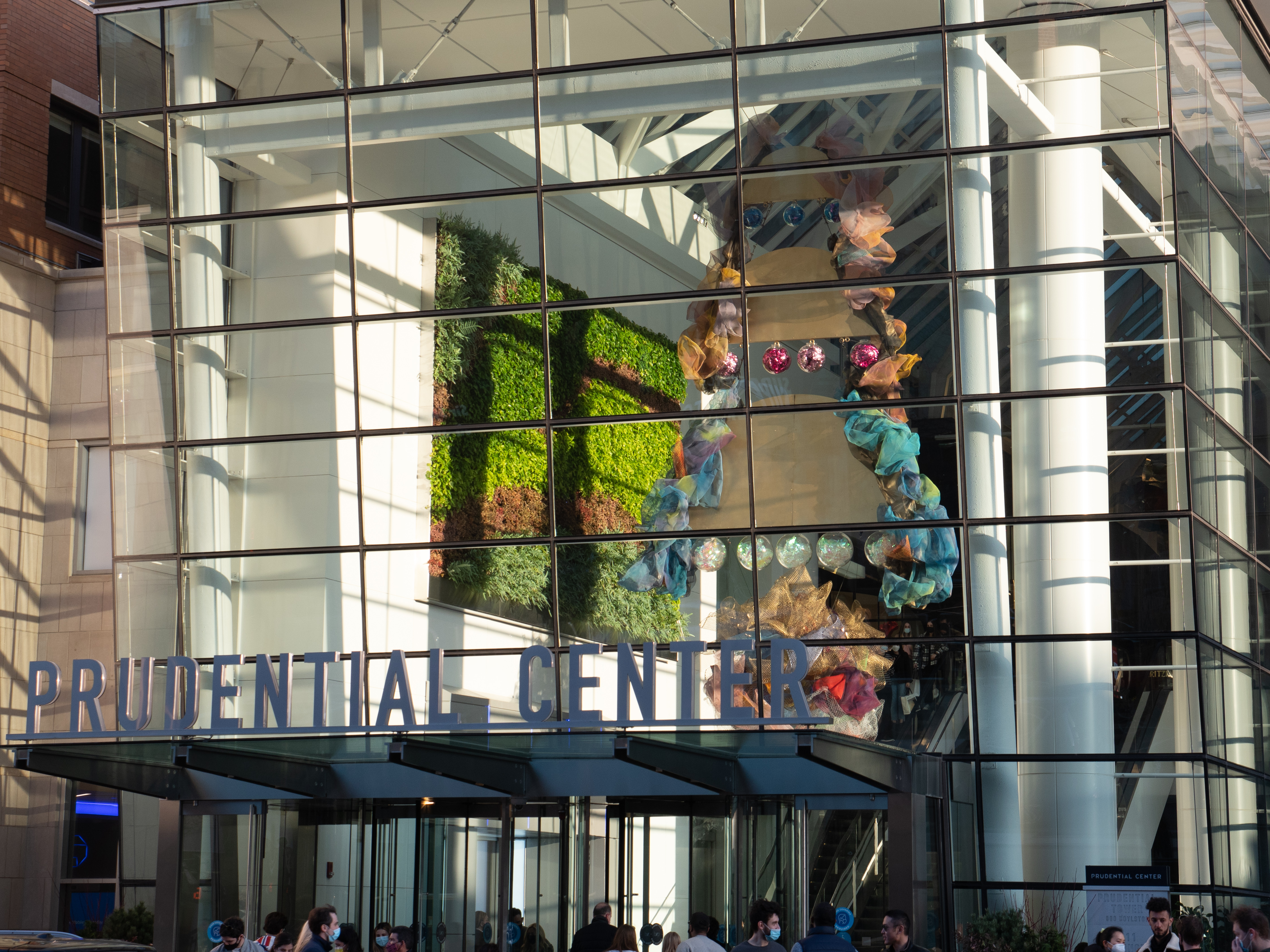 Three half circles with a border of mesh hanging one on top of the other with blue, pink, and translucent mirror balls. A mesh ball hangs below the half circles. The sculpture can be seen through a glass wall behind the "Prudential Center" sign.