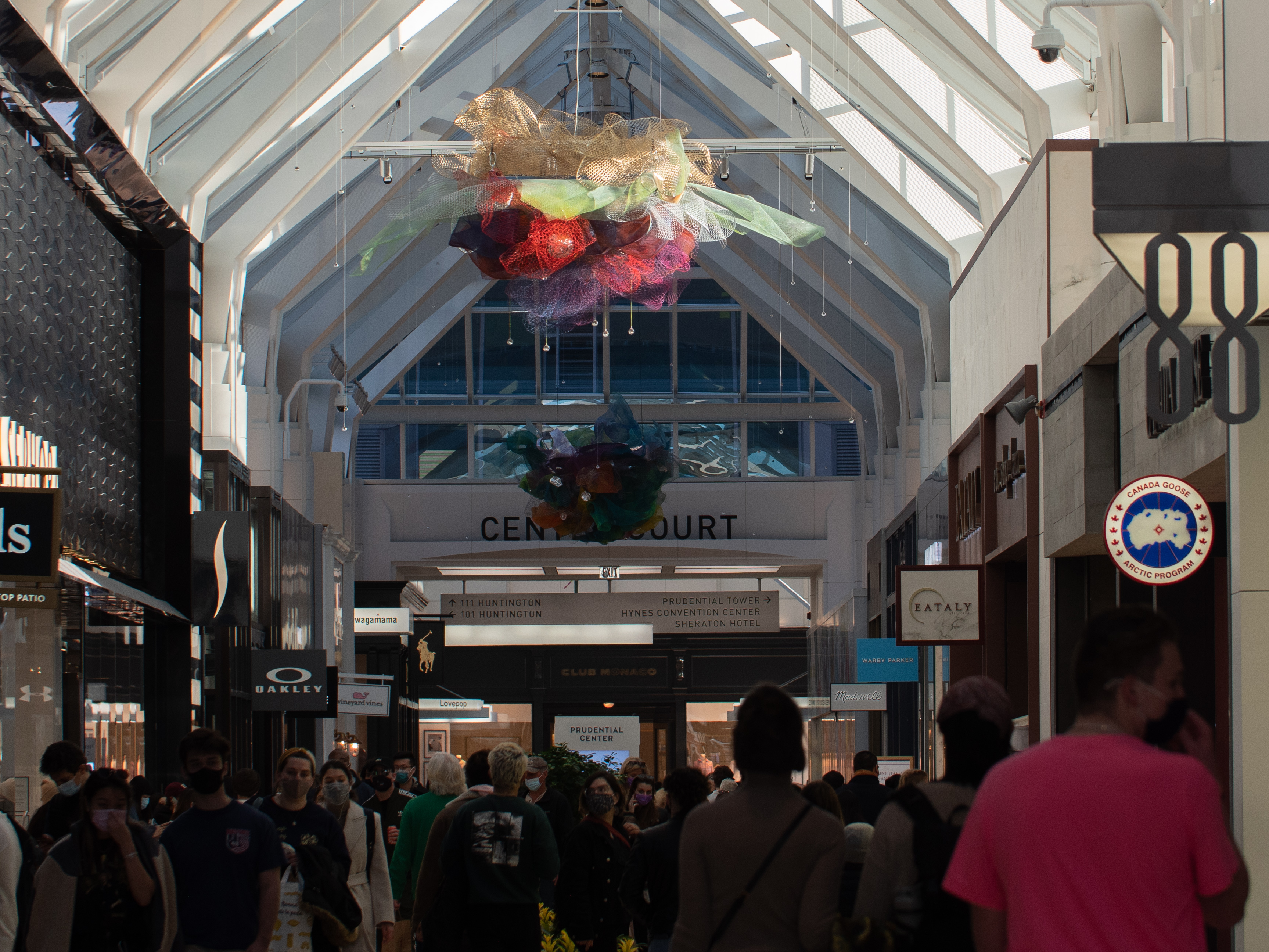 A mesh ball of beige, green, red, purple and a smaller mesh ball beneath of blue, purple, orange, green, and yellow hangs above shoppers and stores in the Prudential Center mall.