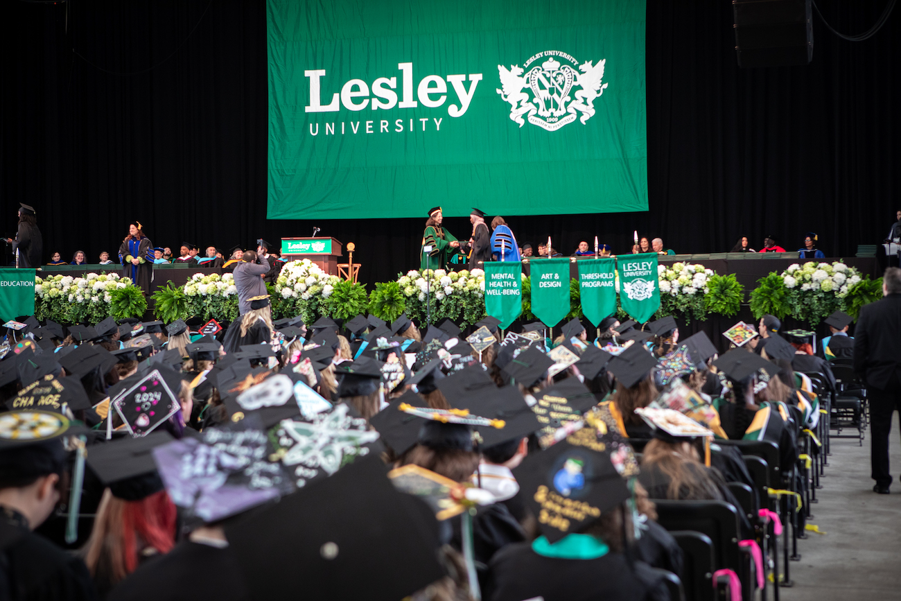 Photo of commencement stage with big green bannister that says Lesley. People sitting with diplomas in chairs facing the stage