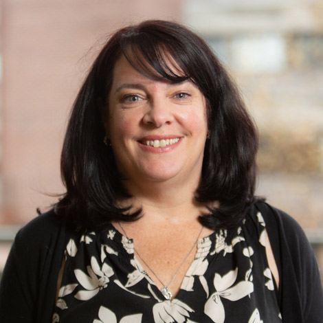 Head shot of counseling center staff member Ellen O'Neill. She is smiling while looking at the camera. She has black hair and wears a black cardigan over a black shirt patterned with white flowers.