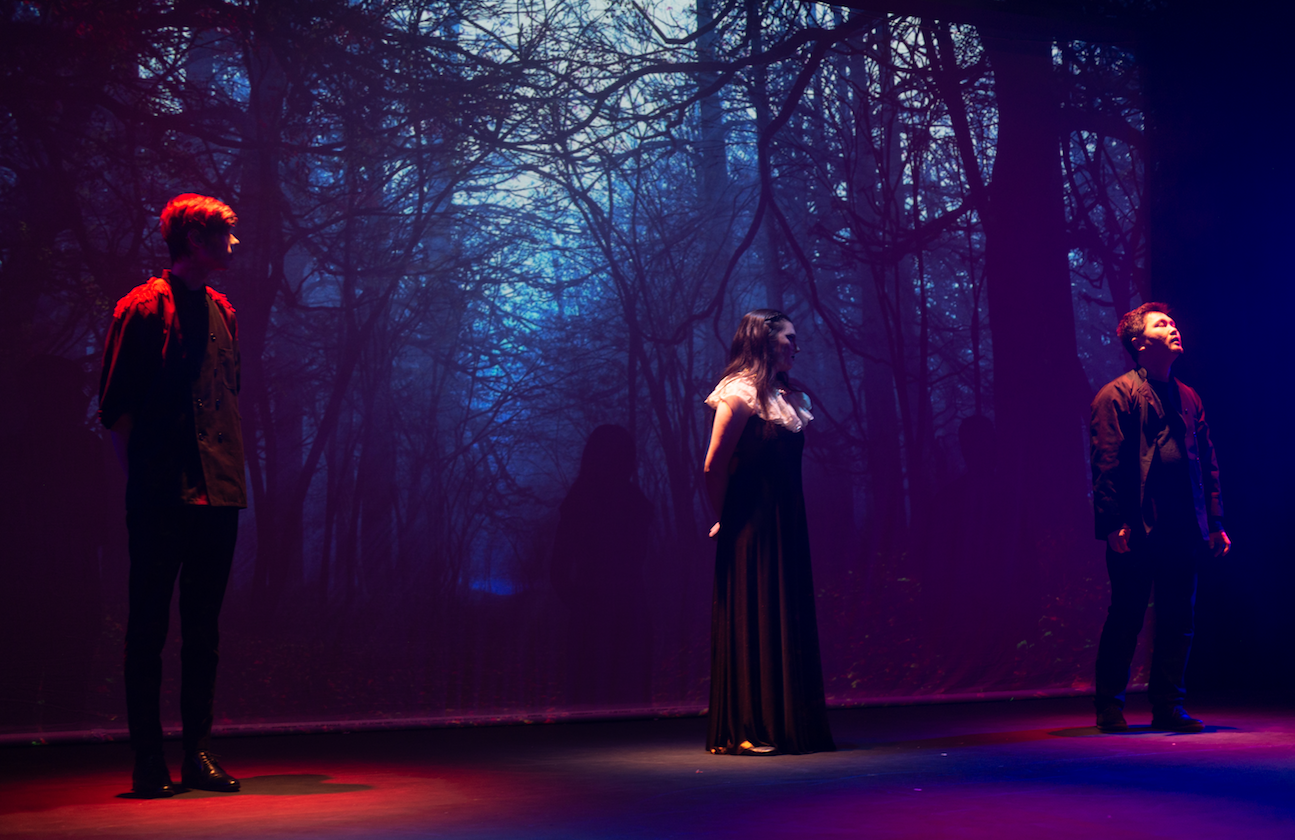 Three actors on stage against the backdrop of the woods in a production of Macbeth