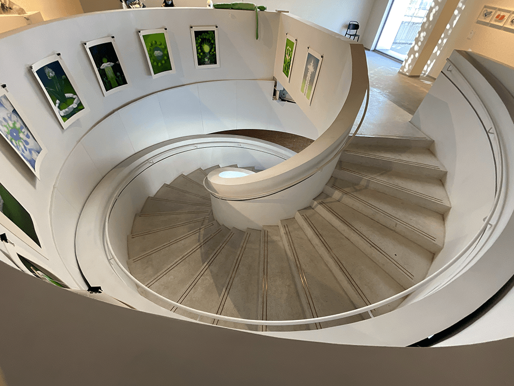 A piece of artwork by Fangwei Xu. It is a white, spiral staircase with artwork on the wall around it.