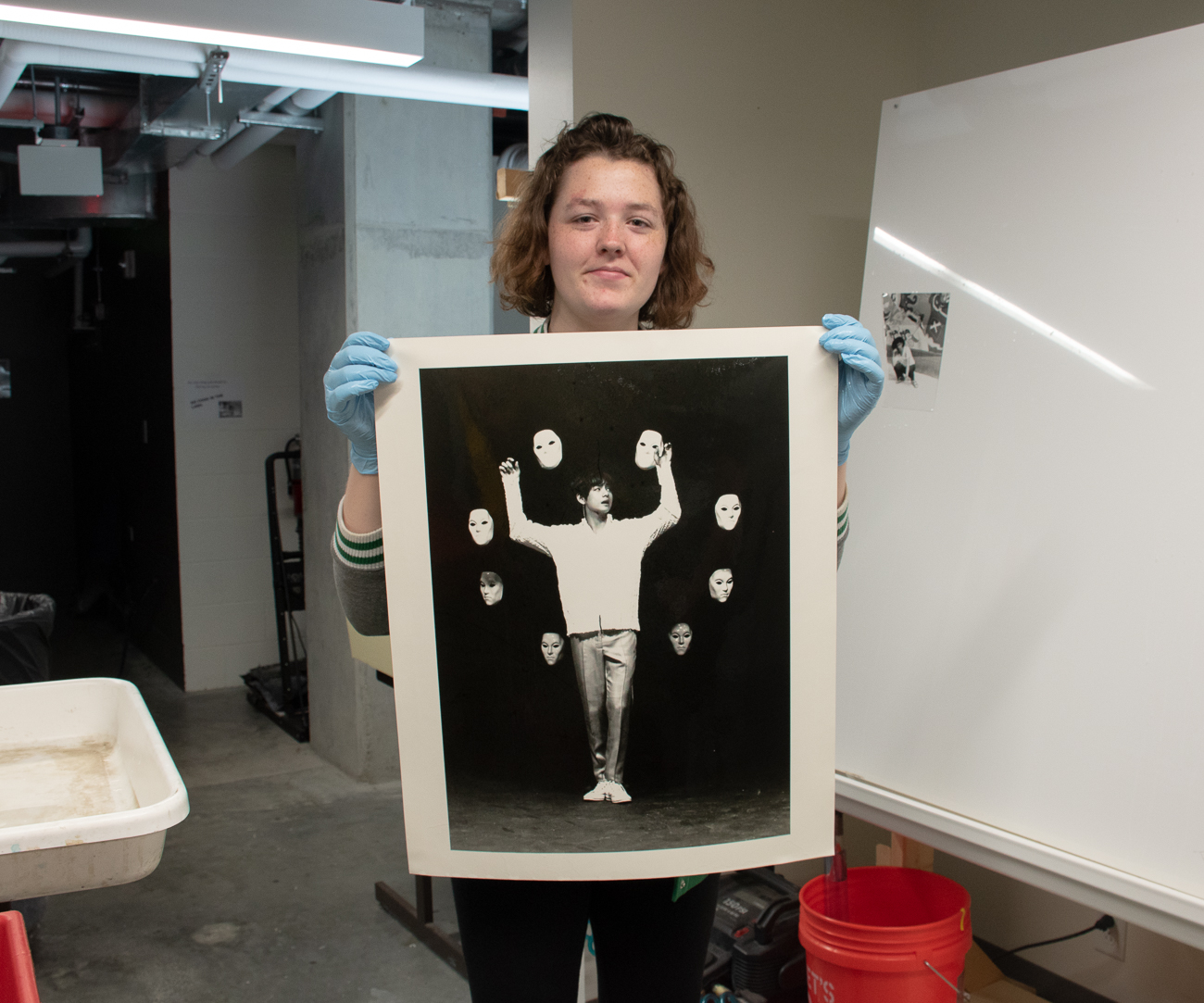 A female student holds up a large photo print of a man with his arms up surrounded by what looks to be porcelein heads against a black backdrop.