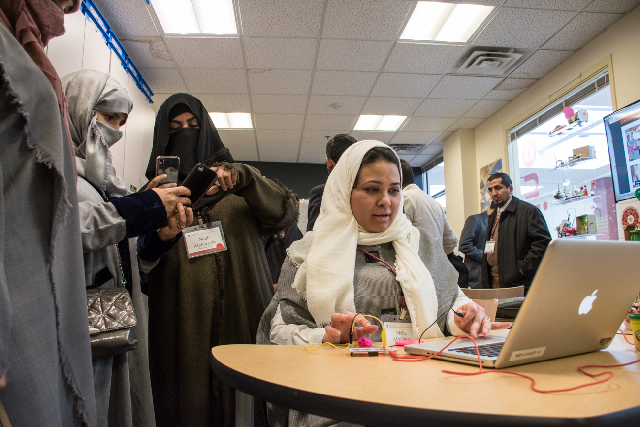 A Saudi woman sits at a computer with other women standing behind her taking photos of her with their phones.