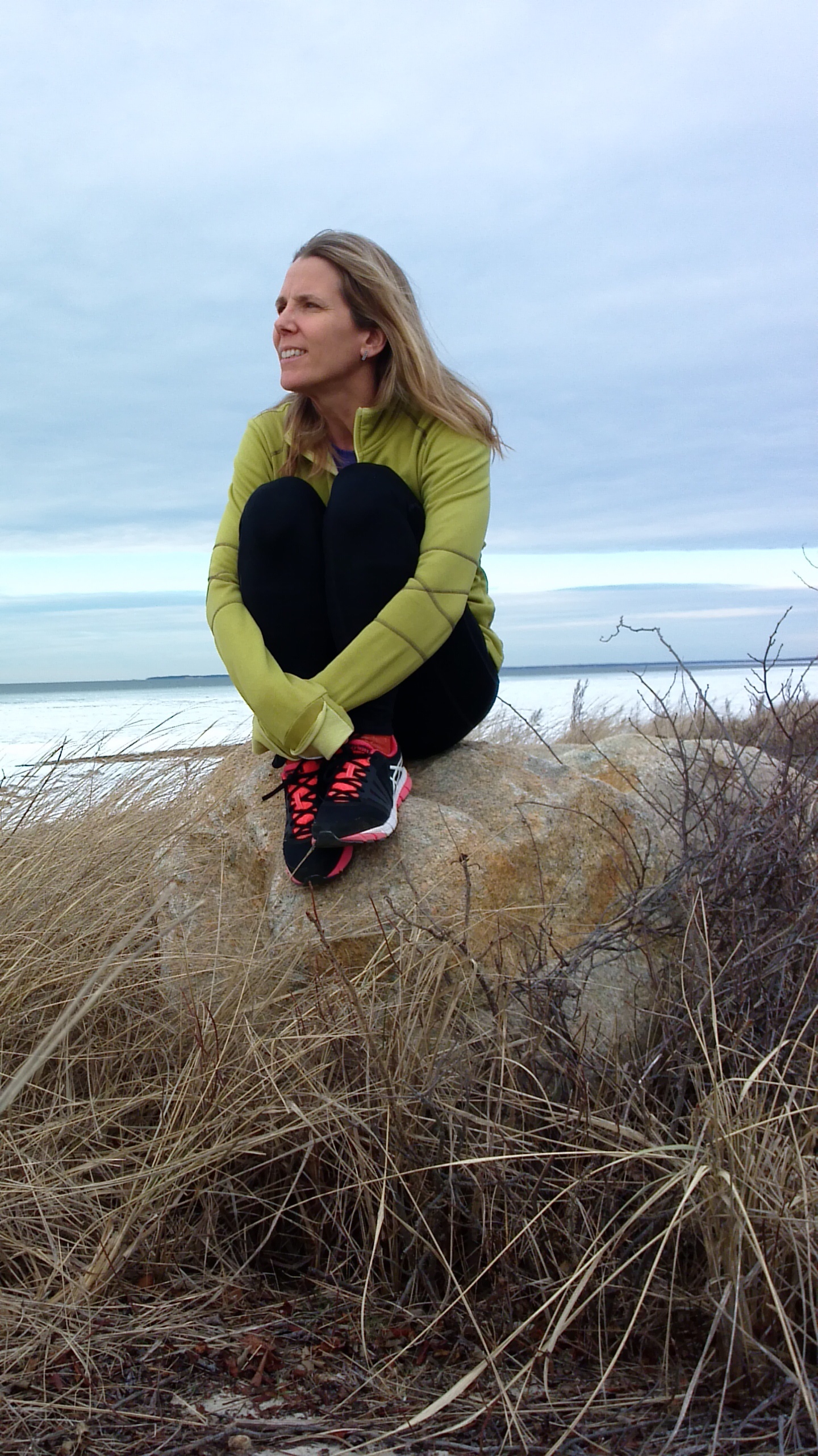 Woman sitting on rock at the beach with long sleeve shirt and pants on, pulling her legs towards her chest and looking off into the distance
