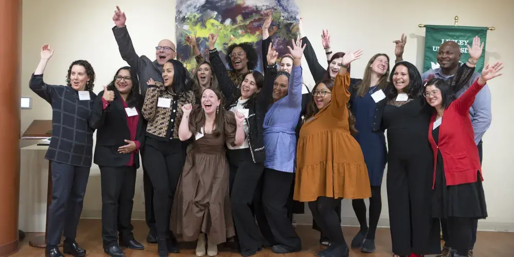 A diverse group of about fifteen professionally dressed adults stand together, posing for a photo. They are all smiling and cheering with excitement, many with their arms raised victoriously. They are indoors in a room with wood floors, a large abstract painting, and a green banner on the wall behind them.