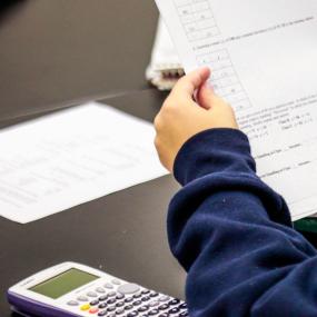 A student sitting at a desk with a calculator and scrap paper holds a worksheet with math questions.