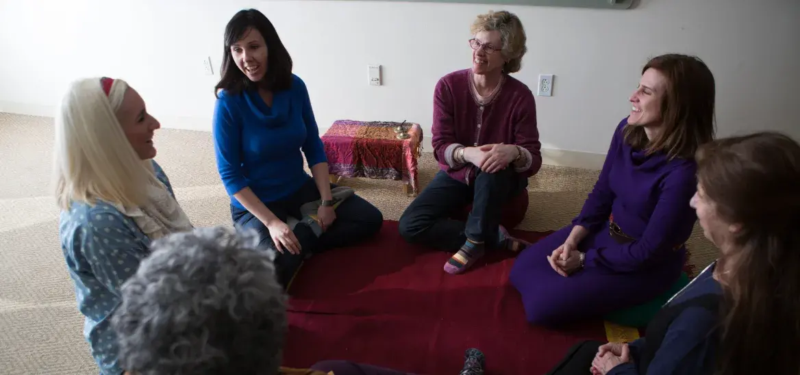A group of six women sits in a circle, with six participants sitting on a red mat on the floor. Their engaged and comfortable postures create a sense of community and supportive conversation in a simple, well-lit room.