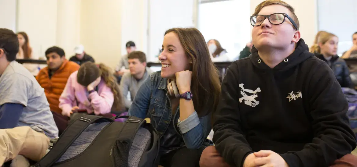 A group of students sit on the floor in a bright, modern classroom. In the foreground, a smiling young woman in a denim jacket and a young man in glasses and a black hoodie appear engaged and relaxed. The atmosphere suggests a collaborative and informal learning environment, with students attentively listening or interacting during a class discussion.