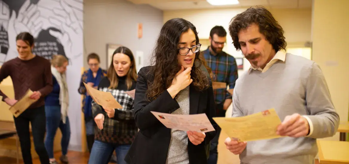 A group of adults stands in a brightly lit hallway or classroom, each holding a sheet of paper and reading intently. Two individuals in the foreground, a woman in glasses and a man in a light sweater, appear to be discussing the content. Others in the background are engaged in similar activities. The setting suggests an interactive or workshop-based learning environment, with casual attire and engaged expressions. A wall mural with hands and text is partially visible in the background.