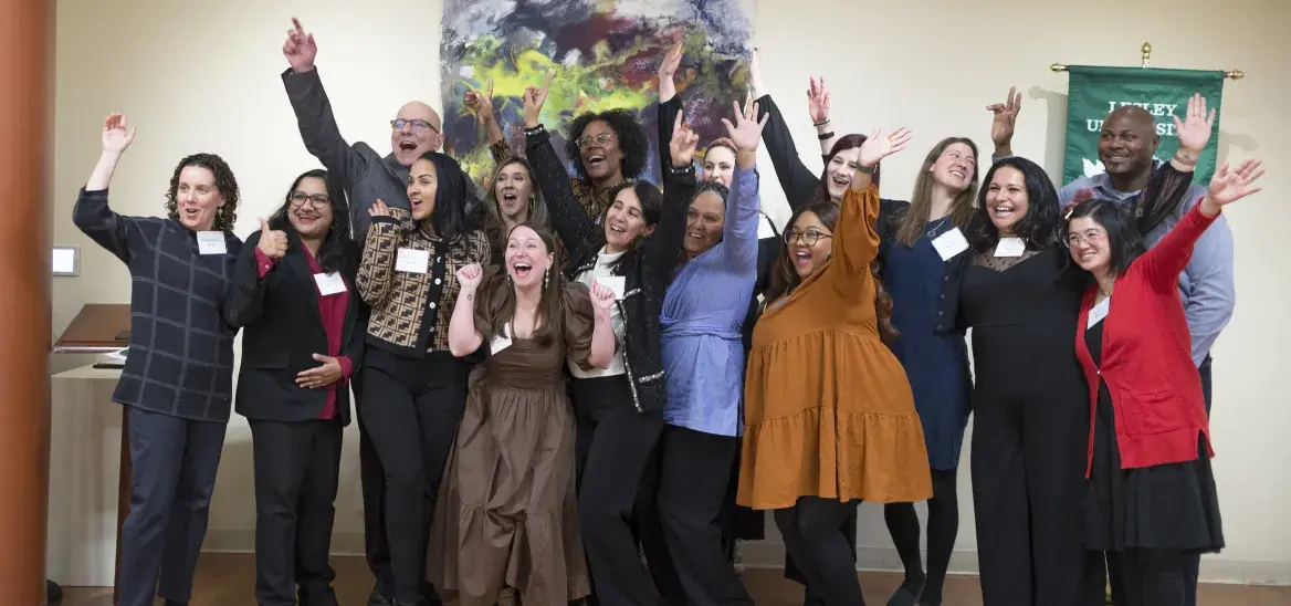 A diverse group of about fifteen professionally dressed adults stand together, posing for a photo. They are all smiling and cheering with excitement, many with their arms raised victoriously. They are indoors in a room with wood floors, a large abstract painting, and a green banner on the wall behind them.