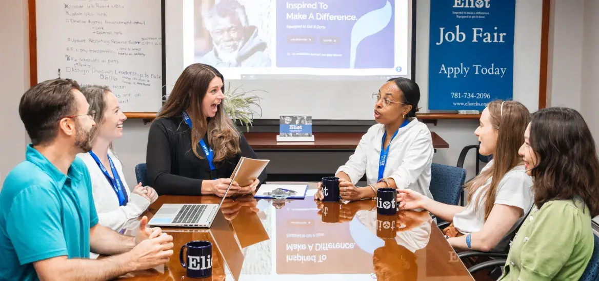 Six people sitting around a meeting table in front of a PowerPoint presentation