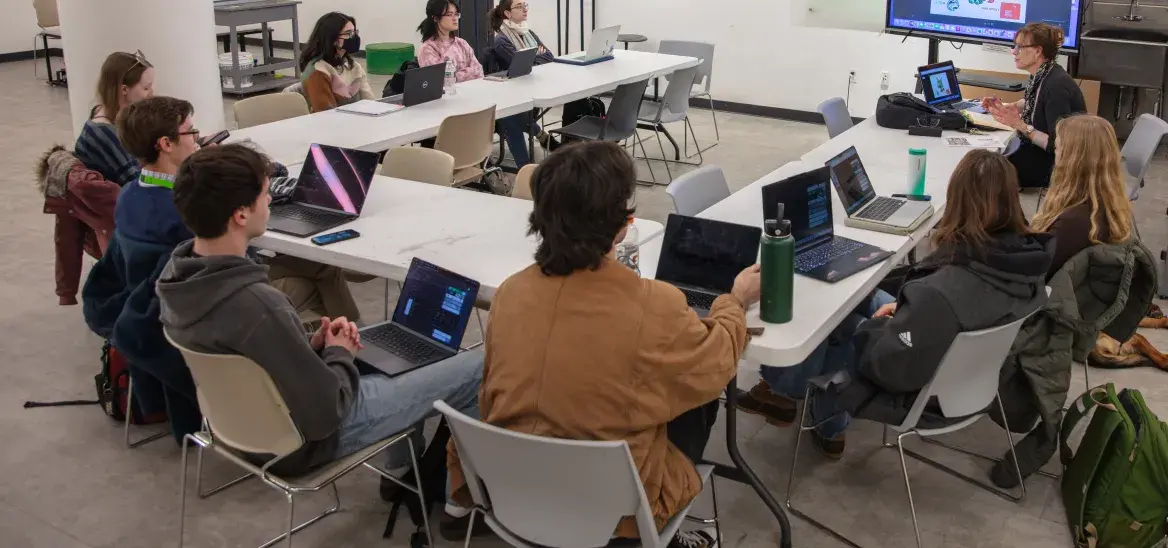 Students around a U-shaped table listening to an instructor in front of an animation screen