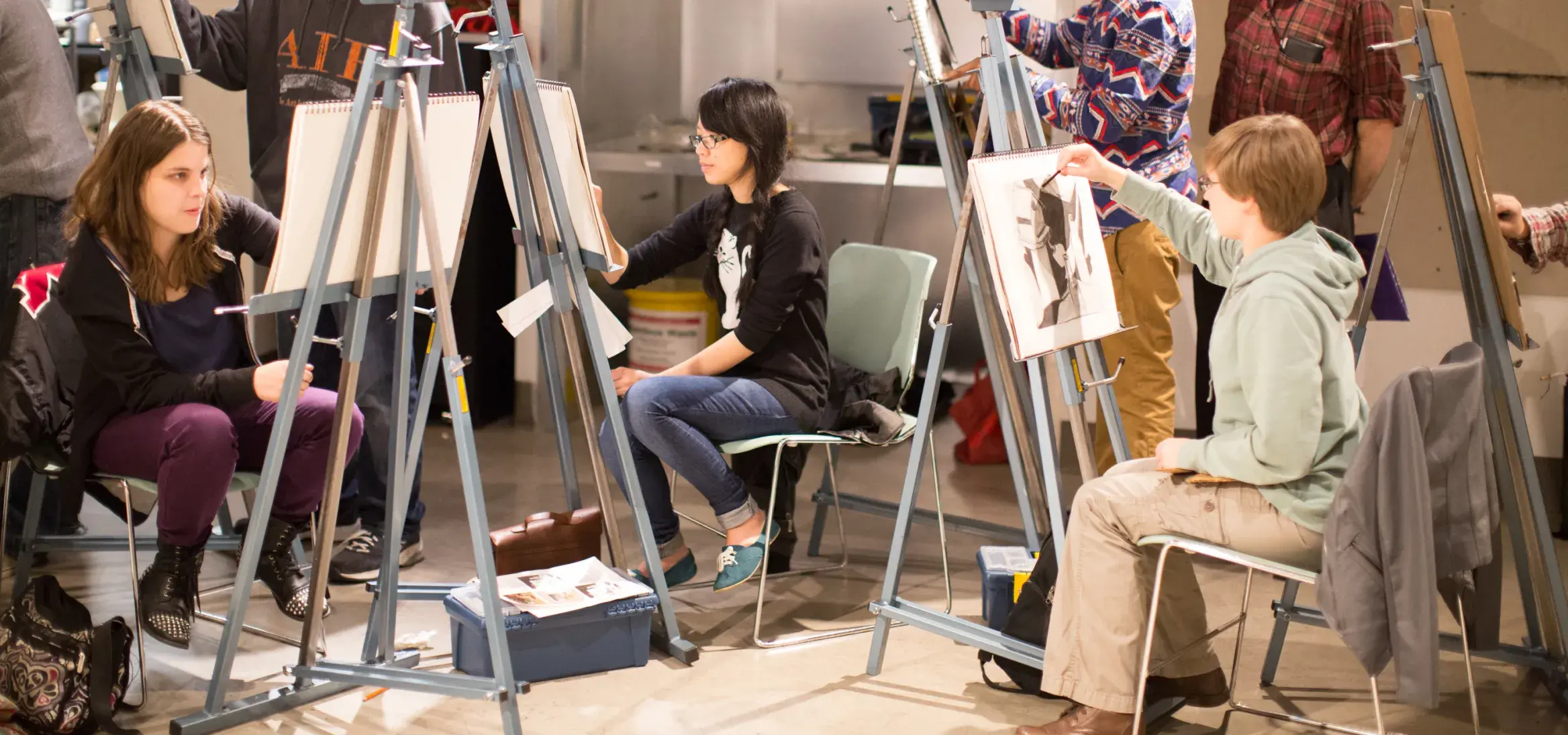 In a busy art studio, a diverse group of students of various ages participates in a life drawing class. They are scattered around the room, drawing on easels. In the foreground, a student holds up their charcoal portrait for critique, while an instructor looks on in the background.