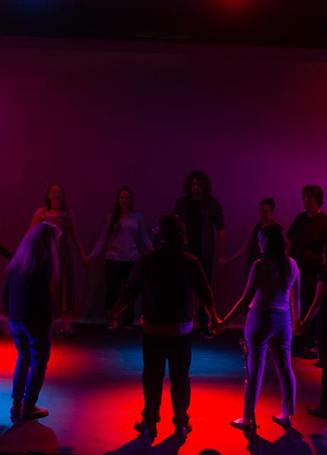 A group of actors in a circle on stage in Marran theater with dark, colorful lighting