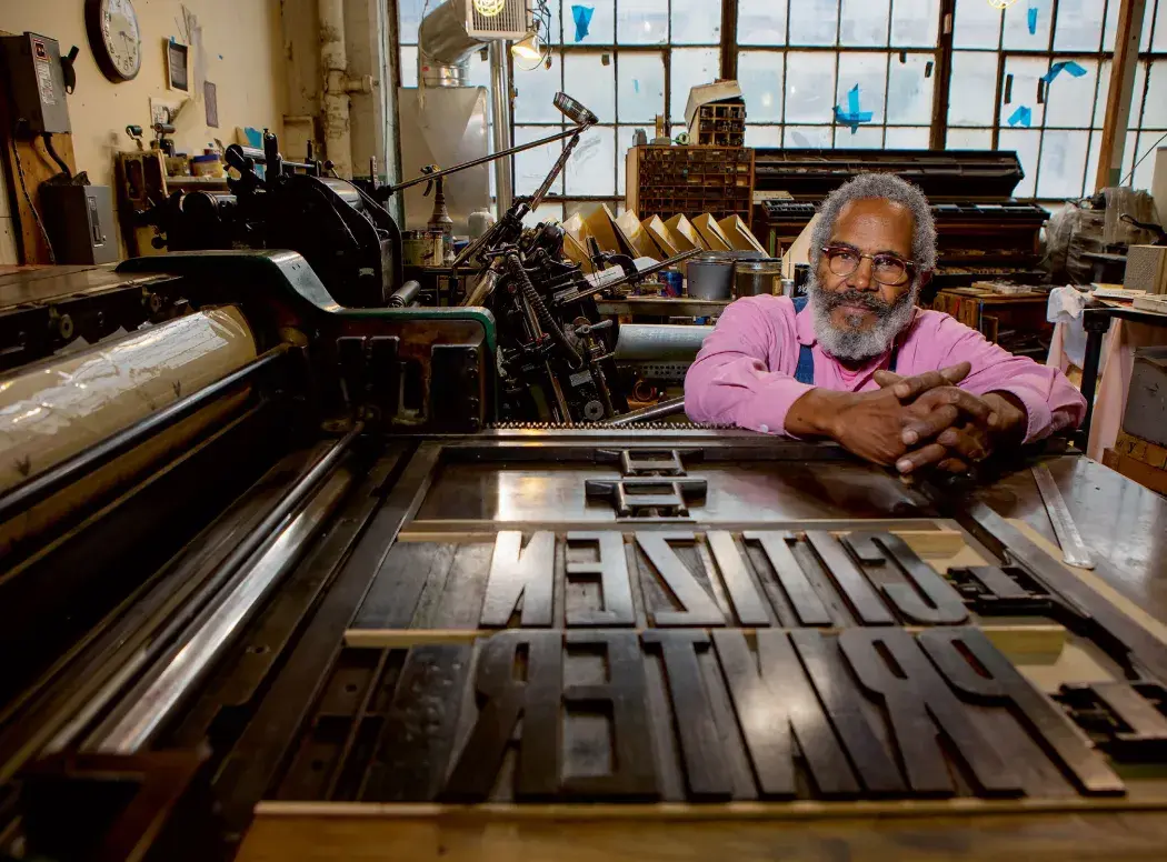 Man sits facing the camera over a large printing press.