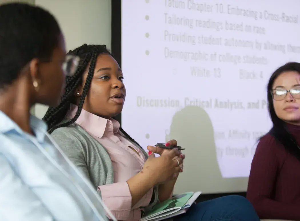 Three students sit speaking in front of a projector screen