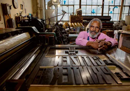 Man sits facing the camera over a large printing press.