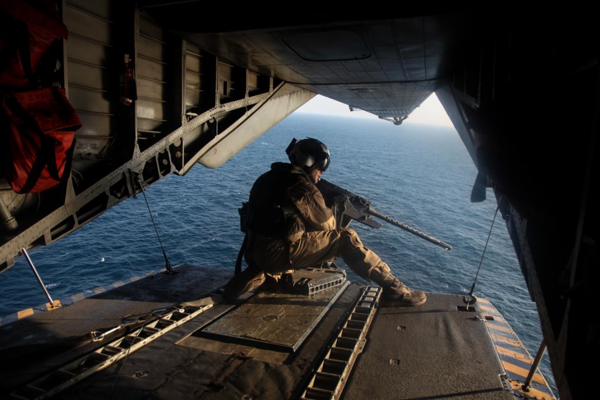 GULF OF OMAN, U.S. Marine Sgt. Gilbert Hopper provides security while flying aboard a CH-53E Super Stallion. Man crouched with gun on edge of open plane back with water in the background.