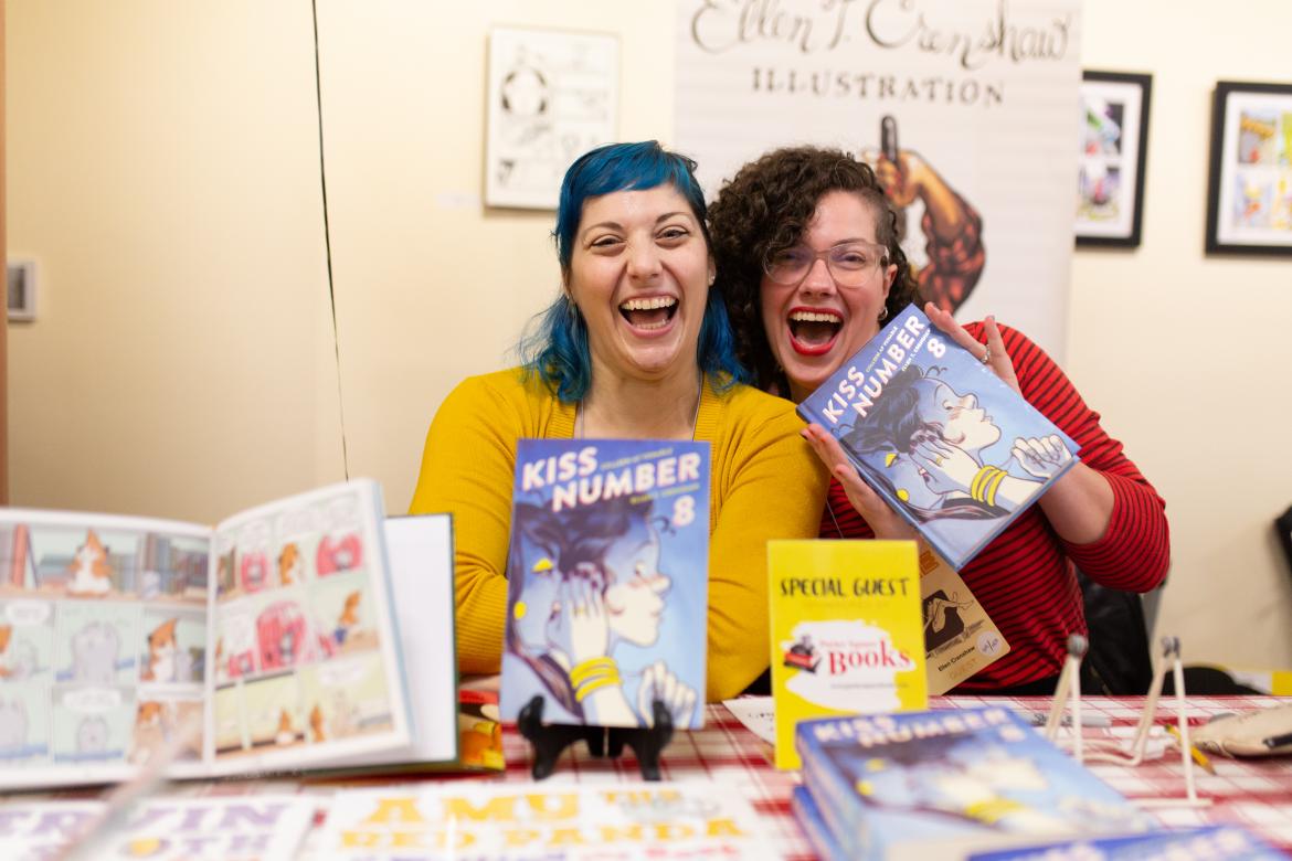 Photo of 2 women behind a table holding up comic books and smiling and laughing