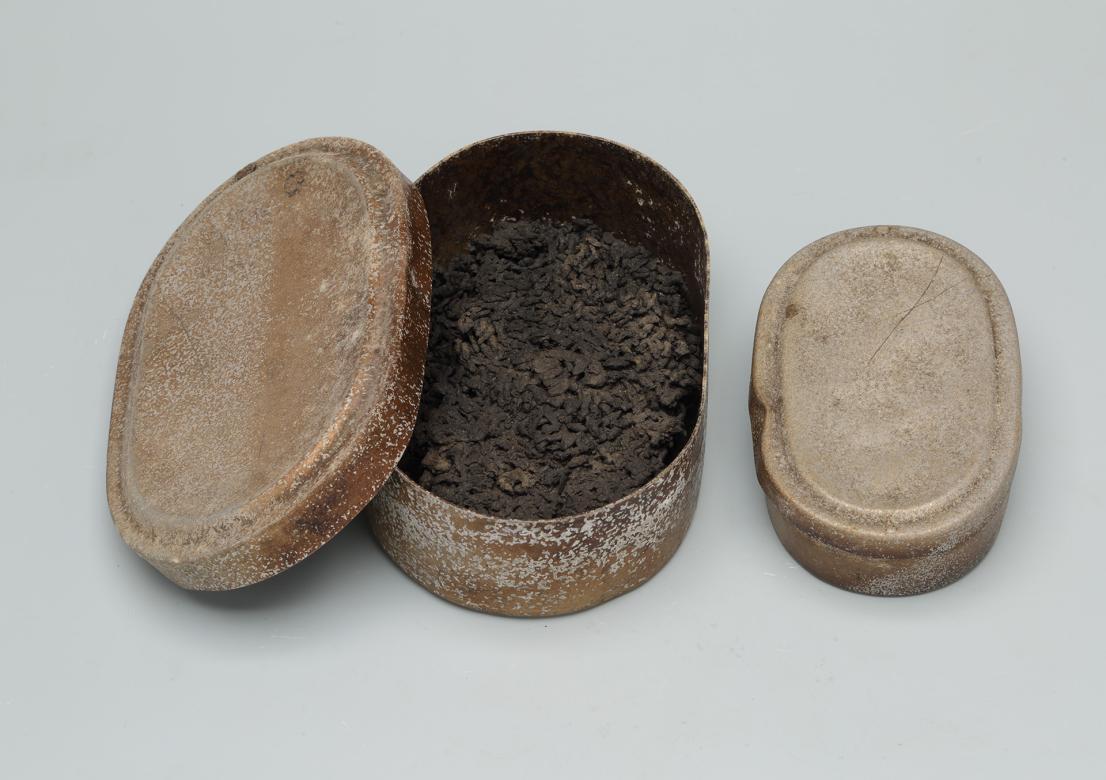 A oval-shaped, light brown metal box/container, with ashes clumped together inside of it.
