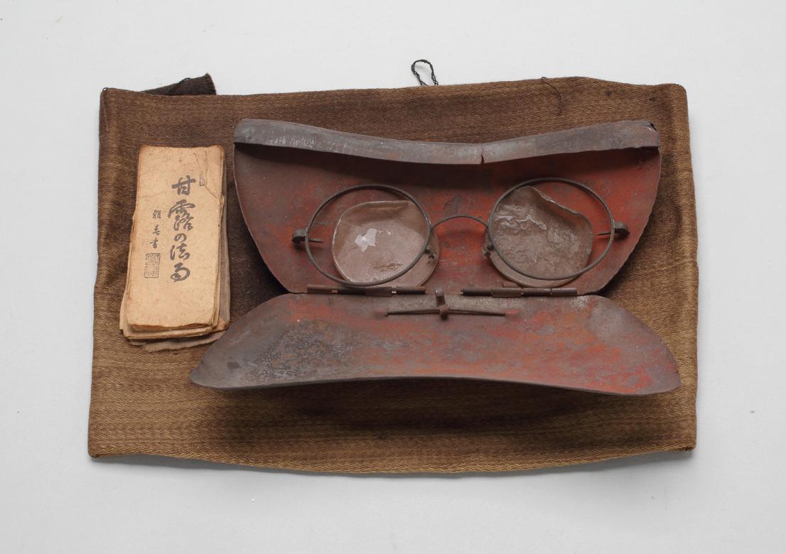 A photograph of damaged glasses inside of a glasses case, which is on top of a small brown pouch.