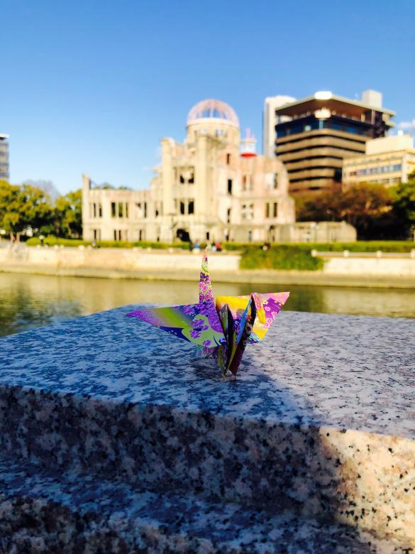 A multicolor paper crane on top of marble steps by a river in Hiroshima during sunrise or sunset.